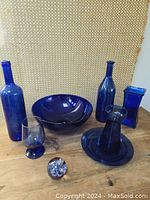Full view of cobalt blue glass assortment on wood surface with textured background, showing two bottles, bowl, candle holder, glass, rectangular vase, and paperweight.
