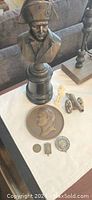 Photo showing the bronze-like napoleon bust on black pedestal along with two brass cannon miniatures and various historic coins and medals on white cloth.