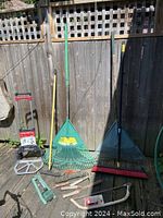Photo showing the full assortment of garden tools and hand tools leaning against a wooden fence on deck, including leaf rakes, push broom, and hand tools.