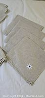 Set of six beige linen napkins with embroidered daisy on each, laid out on white tablecloth.