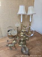 Photo showing pair of brass candlestick electric lamps with white shades, two brass flower-shaped candle holders, small brass-bound wooden barrel, brass figurines of unicorn and horse, and a small brass candle holder.