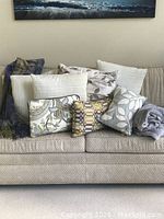 Nine assorted throw pillows and two blankets arranged on a beige sofa. Pillows include leafy, geometric, and white textured prints. Two blankets, one gray and one plaid chenille, are visible.