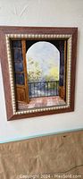 Framed painting showing an arched doorway with black rail and garden beyond, positioned on light textured wall.