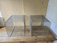 Two chrome wire cubes side by side on wooden surface with woven backdrop