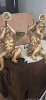 Pair of gold painted classical style angels with trumpets, positioned kneeling on wooden furniture surface.