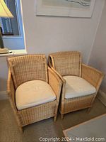 Pair of wicker rattan chairs with cream cushions placed side by side indoors under lamp light