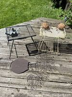 Wide photo showing two metal patio tables, one black and one white, with mesh tops, a black and white house watering can, and three pieces of hanging metal garden decor including a vase and two sun face shapes.