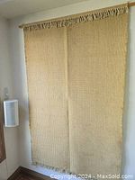 Full view of wall hanging mounted on wall showing overall size and tassel detail. Natural beige color with woven texture.