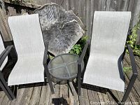 Two grey mesh fabric patio chairs with dark metal frames and two round glass top tables with metal legs placed outdoors on wooden deck.