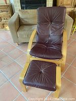 Front view of chair and footstool showing reddish brown pebble grain leather cushions with button details and birch bentwood curved frames, placed on tiled floor.