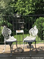 Two white wrought iron patio chairs and a small wrought iron side table on wooden deck with greenery background.