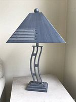Front view of vintage postmodern table lamp with rectangular openwork wavy metal body, rectangular tapered shade with cutout horizontal lines, and ball finial on top. Matte pewter textured finish.