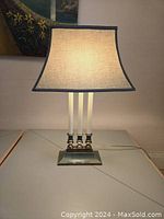 Front view of the vintage table lamp illuminated, showing three white columns on a rectangular green base and a curved rectangular shade with dark trim.