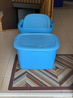 Four blue plastic storage bins stacked, with lids on top, displayed on a patterned rug near stairs.