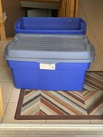 Front view of 2 stacked blue plastic storage bins with gray lids on a patterned rug in a tiled area.