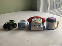 All four items displayed on a bed: Fisher Price rolling telephone, ceramic truck mug, Contigo stainless steel travel mug with lid, green monster truck toy.