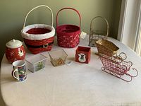 Photo showing all baskets and ceramic items arranged on a table: large Santa basket, large candy cane basket, medium wire basket, two sleigh-shaped wire baskets, three small metal baskets, ceramic cookie jar, and mug.