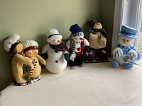 Group photo of five Christmas stuffed decorations lined up on a shelf against a green wall.