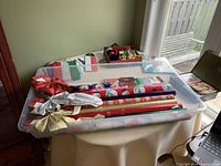 Photo showing plastic container filled with assorted Christmas wrapping supplies including wrapping paper rolls, bows, ribbons, cards, invitations, tape, cutter, and tags.