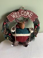 Front view of a decorative grapevine wreath with artificial evergreen, pine cones, berries, a plush moose figure in blue sweater and red pants holding ski poles, and a maroon wooden welcome sign at top.