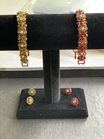Two gold-toned bracelets displayed on a black stand with matching pairs of earrings below; one bracelet has green leaf-shaped and pearl details, the other has red and pearl accents.