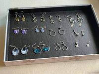 12 pairs of silver tone earrings arranged in a box with black background showing enamel earrings, hoops, and studs