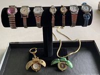 Nine items displayed on a black tray and black watch holder stand. Includes seven wristwatches with various face and band styles, a ladybug keychain watch, and a ladybug pendant necklace.