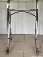 Front view of silver walker showing adjustable legs with height holes, black and red plastic parts, and two front wheels with rubber caps on rear legs.