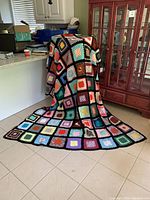 Full view of the king size crochet blanket draped over a chair and extending onto the floor, showing colorful squares bordered in black.
