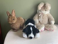 Group photo showing all three stuffed animals: large beige bunny, medium brown bunny, and medium black and white puppy seated on a wooden surface