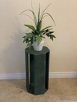 Full view of octagonal dark green wooden plant stand with artificial potted greenery on top, against beige wall and beige carpet floor.