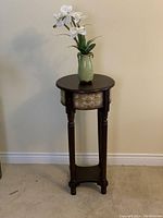 Tall brown wooden plant stand with round top, lower shelf, carved legs, and fabric floral skirt with green vase and white faux orchids on top.