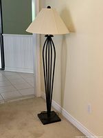 Tall floor lamp with black metal base featuring vertical curved rods and a white fabric shade, shown standing on carpeted floor near a cream wall and pink armchair in a living space.