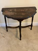 Side view of half-moon shaped wooden table with three turned legs and scalloped edges showing wear on surface.