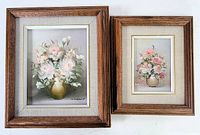 Two small floral oil paintings with wood frames shown together, depicting pastel flowers in vases with beige fabric mats and gold trim.