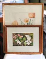 Two framed floral watercolour paintings stacked with visible wooden frames; larger with peach tulips, smaller with white/pink flowers.