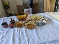Photo showing a variety of amber glass bowls, including one large bowl and smaller bowls, along with several small ceramic and glass decorative items arranged on a white cloth.
