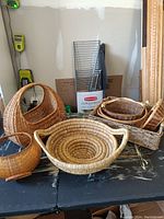 Seven assorted wicker baskets in various shapes and weaving styles displayed on a black table.