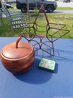 Photo showing wooden apple-shaped ice bucket, star-shaped metal wine rack, and tin box with decorative lid on a blue cloth background outdoors.