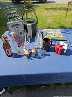 Full display of vintage barware items on blue tablecloth outdoors including ice bucket, poker chips, glass pitcher, bottle openers, coasters and coffee cup.