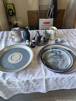 Overall view of the ceramic pottery lot including trays, oil and vinegar set, mugs, and small decorative pieces on white tablecloth