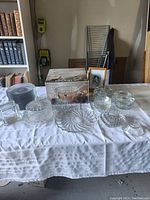 Full view of all clear glass items on a white tablecloth including plates, punch bowl box, cake keeper, and stacking bowls.