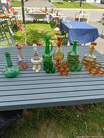 Full view of all decanters and matching glasses arranged on a patio table showing various coloured glass decanters with green, amber, and clear hues, some with painted sailboats.
