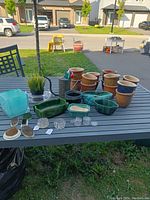 Overview of vintage gardening lot with various ceramic pots, glass flower frogs, green glazed planters, metal watering can with fake flowers, and other items on a table outdoors.