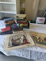 Photo showing assorted hardcover books stacked along with two Egyptian papyrus prints on a table.