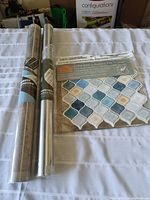 Three home renovation adhesive products for wall covering, including two wallpaper rolls (silver metallic and tile pattern) and a pack of self-adhesive tile stickers in blue and beige tones.
