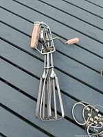 Vintage metal hand mixer with wooden handle and crank resting on grey surface, showing beaters and turning mechanism