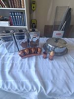 Photo showing full lot items including four metal canisters, wooden spice rack with five spice containers, two wooden egg cups, wooden egg, large turned wood candle holder, and metal cake tin.