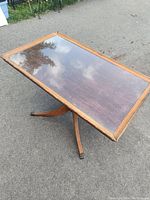 Rectangular wooden occasional table with removable glass top and brass capped feet, standing outdoors on pavement, showing angled side view with pedestal base and legs.