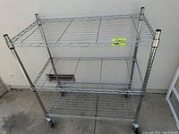 Front and side angled view of 3-shelf metal wire shelving unit with wheels, placed outdoors near a beige wall and door.